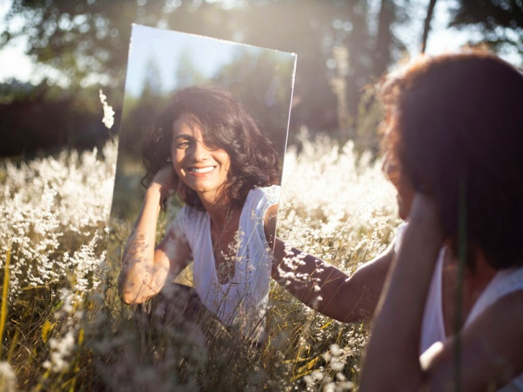 Γυναίκα καθισμένη στη φύση κρατάει έναν καθρέφτη, κοιτάζεται και χαμογελάει.