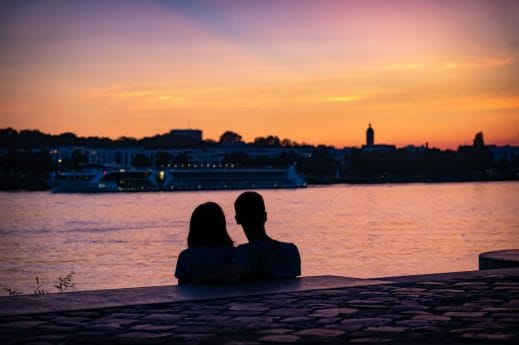 Ζευγάρι (άνδρας και γυναίκα) που κάθεται μπροστά στη θάλασσα και κοιτάζει προς αυτήν με φόντο τον ουρανό σε χρώματα του ηλιοβασιλέματος.