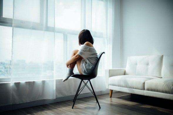 Γυναίκα καθισμένη σε καρέκλα με τα πόδια μαζεμένα μπροστά της κοιτάζει το παράθυρο σε ένα άδειο δωμάτιο