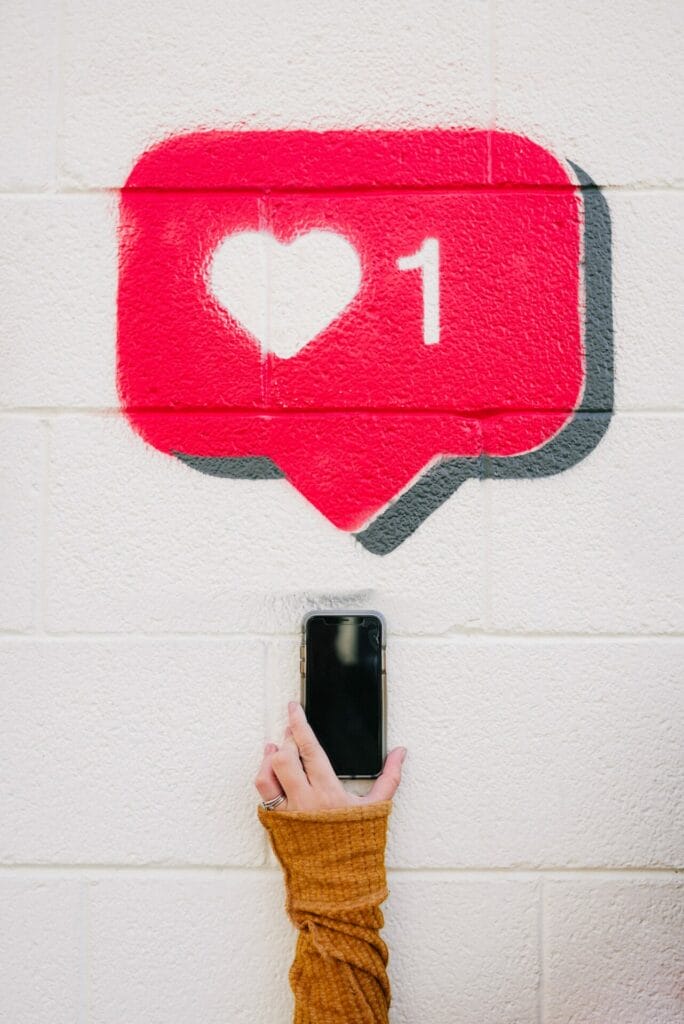 Ένα χέρι που κρατάει ένα smartphone και από πάνω ζωγραφισμένο στον τοίχο ένα like του Instagram