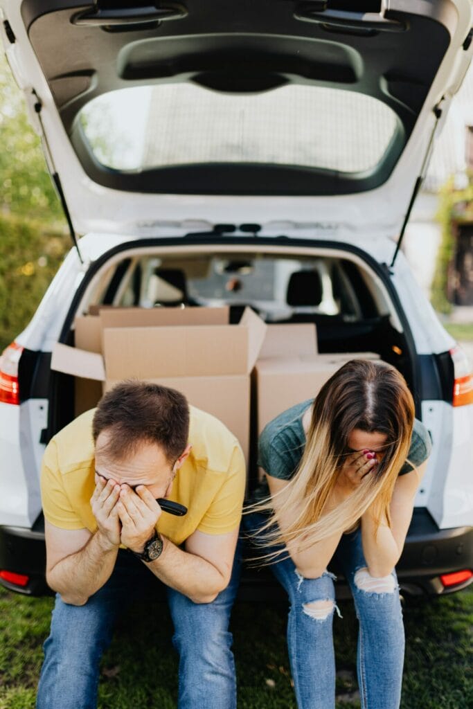 Άνδρας και γυναίκα προβληματισμένοι καθισμένοι στο καπό του αυτοκινήτου που είναι γεμάτο κούτες