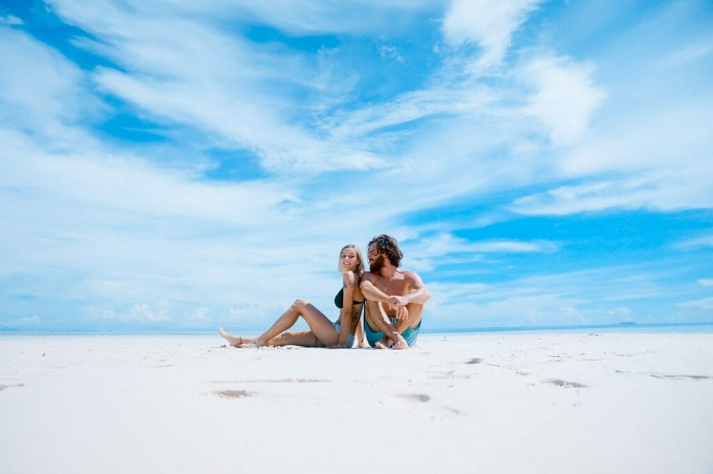Άνδρας και γυναίκα καθισμένοι στην άμμο στην παραλία χαμογελούν.