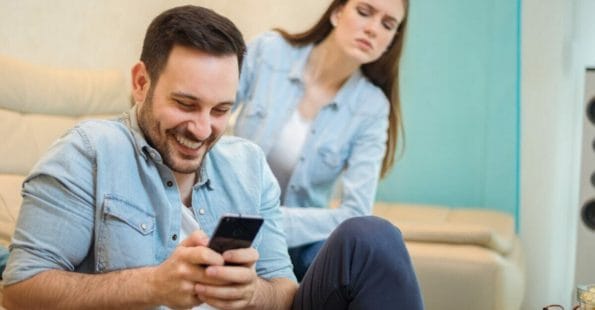 Άνδρας χαμογελάει ενώ ασχολείται με το κινητό του και γυναίκα κοιτάζει προς το μέρος του με βλέμμα ζήλιας